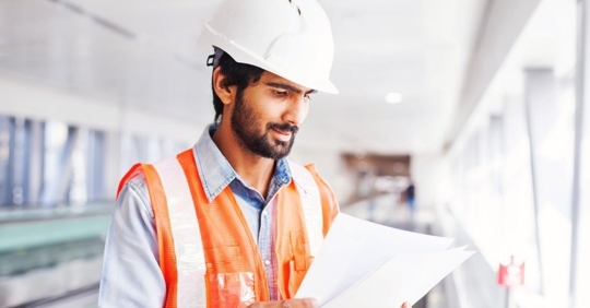 bearded man wearing construction gear (hard hat, reflective vest) looking at papers