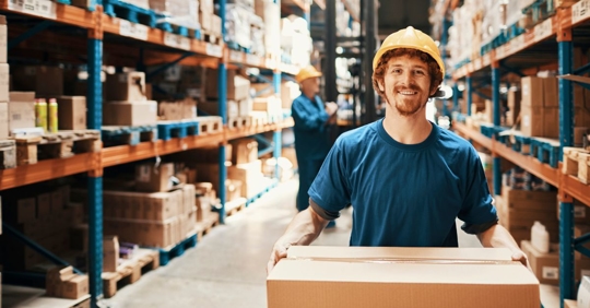 young man wearing a hard hat standing in the aisle of a warehouse smiling and holding a cardboard box