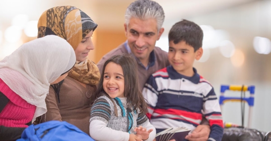 young family sitting together at an airport smiling, there are two woman wearing hijabs and an older man with a young boy on his lap, and a young girl is sitting on an older woman's lap